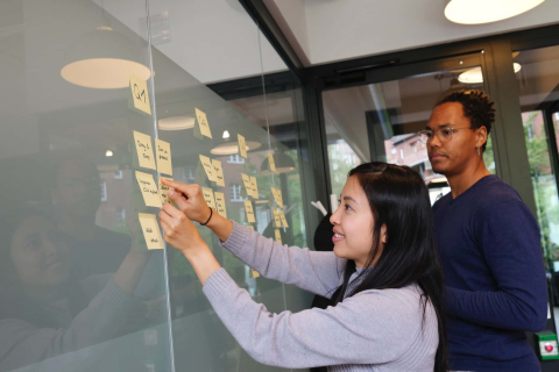 Asian woman and African American man reviewing sticky notes on a wall.